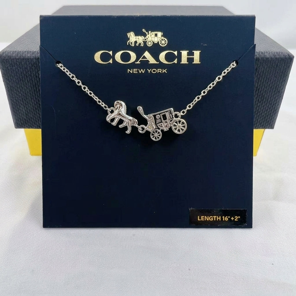 A silver-tone Coach New York necklace with a horse-drawn carriage pendant, displayed on its dark navy blue branded box.