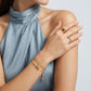 A close-up of a model wearing delicate gold jewelry, including a multi-strand chain bracelet with a small charm, a ring, and a bangle, paired with a light blue high-neck top against a neutral gray background.