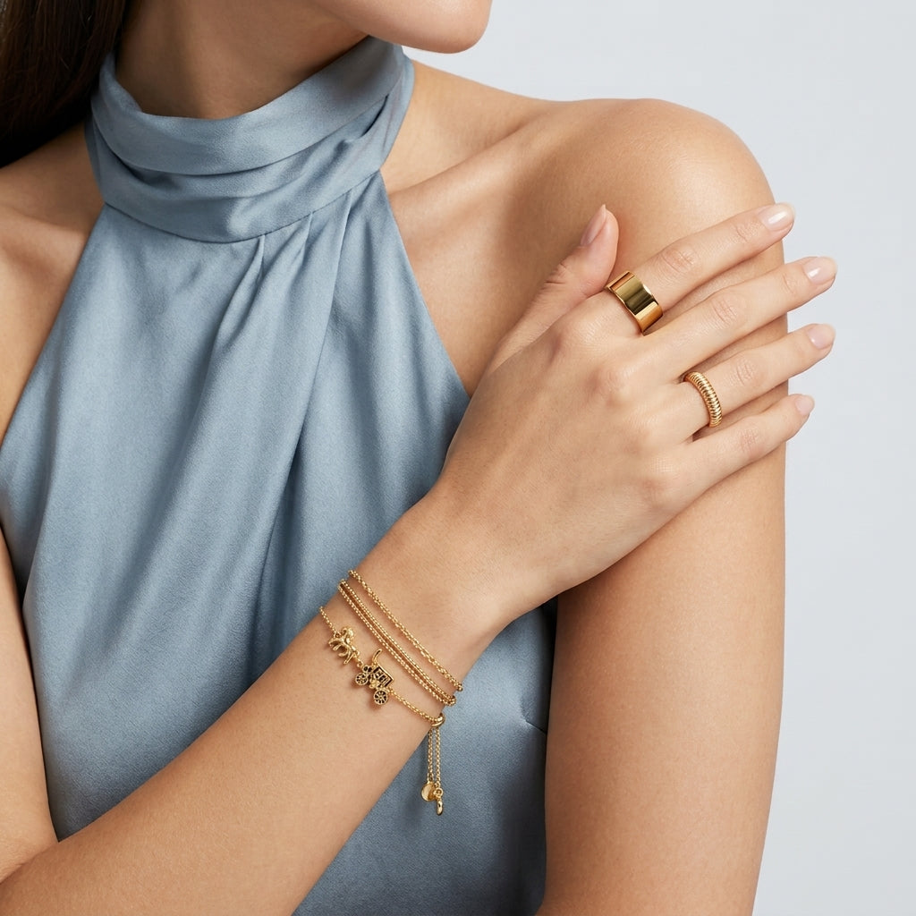 A close-up of a model wearing delicate gold jewelry, including a multi-strand chain bracelet with a small charm, a ring, and a bangle, paired with a light blue high-neck top against a neutral gray background.