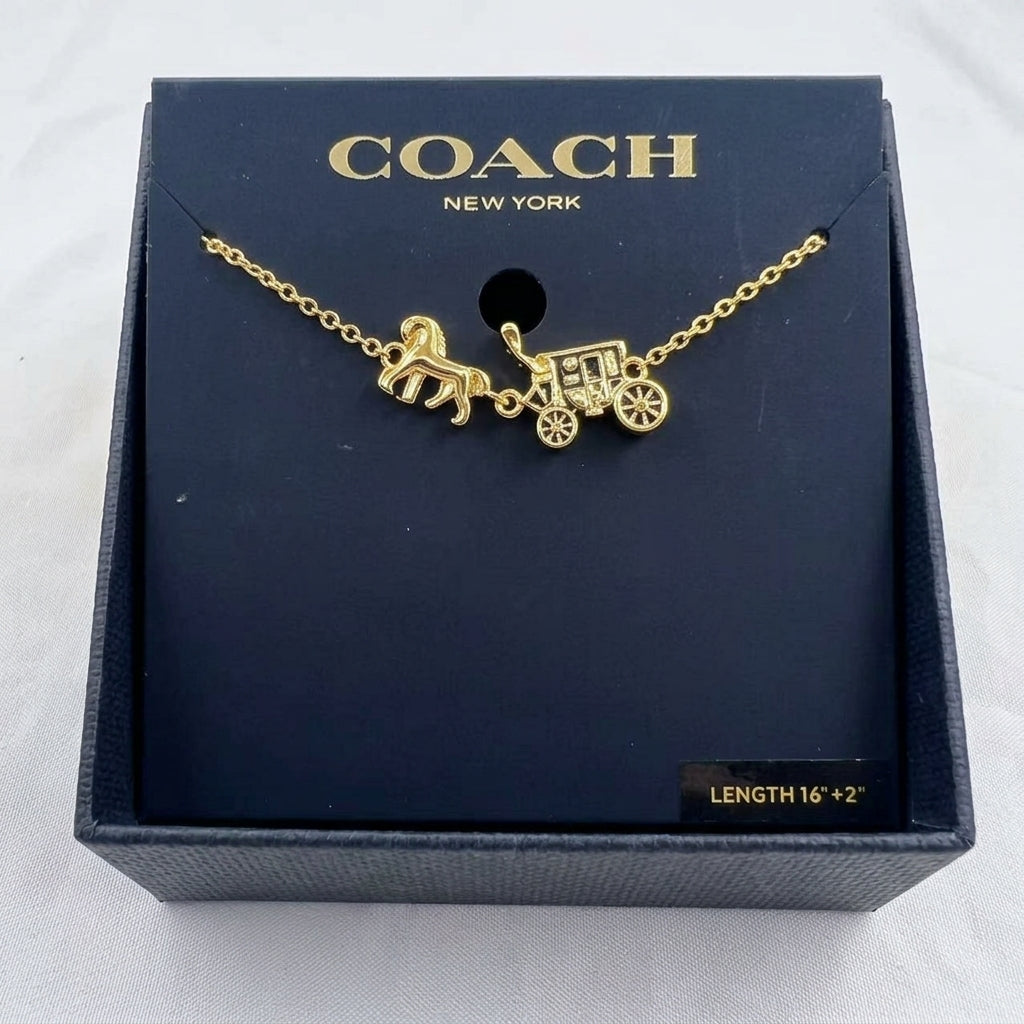 A gold-tone Coach necklace with a horse-drawn carriage charm, presented on a dark navy blue branded display card inside a textured Coach gift box.