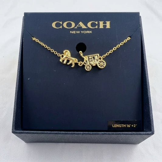 A gold-tone Coach necklace with a horse-drawn carriage charm, presented on a dark navy blue branded display card inside a textured Coach gift box.