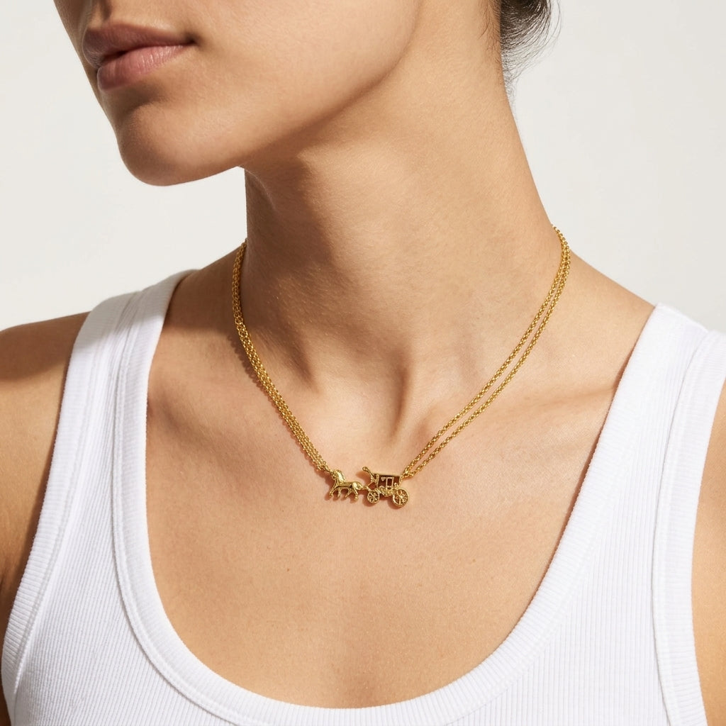 A close-up of a model wearing a delicate gold chain necklace with a small horse-drawn carriage pendant, styled with a white ribbed tank top against a clean, light beige background.