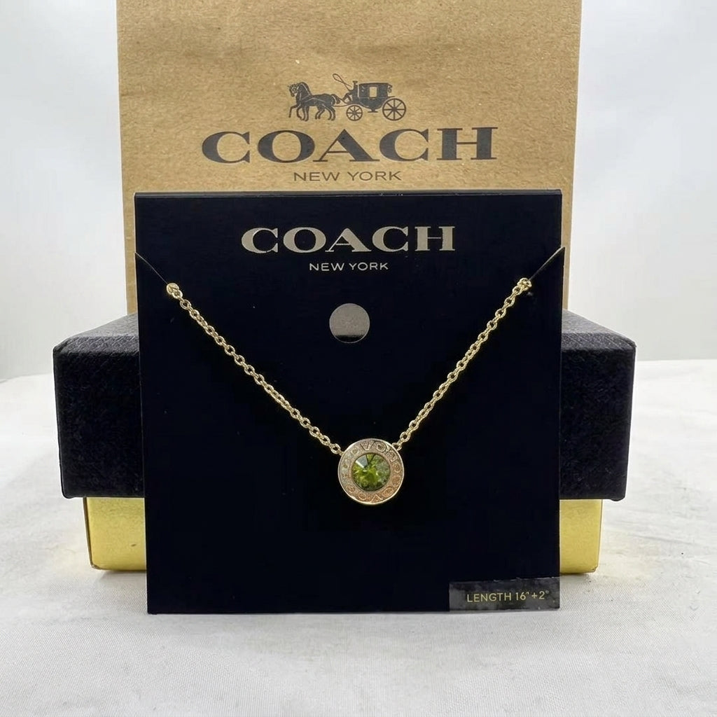 A gold-tone Coach necklace with a round green gemstone pendant, displayed on a black Coach New York card in front of a brown Coach box.