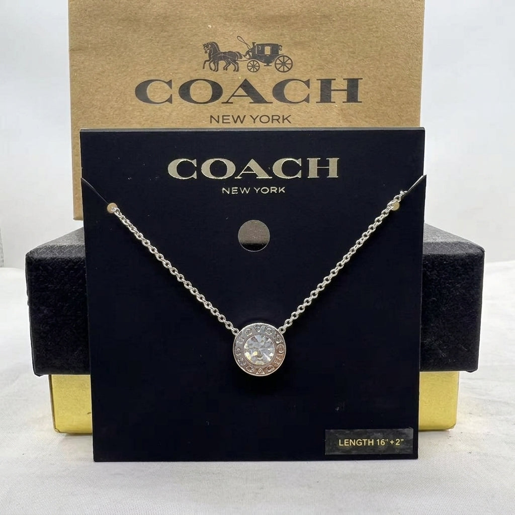 A Coach New York necklace displayed on a black branded card, with a gold-tone box behind it. The necklace has a silver-tone chain and a round clear gemstone pendant.