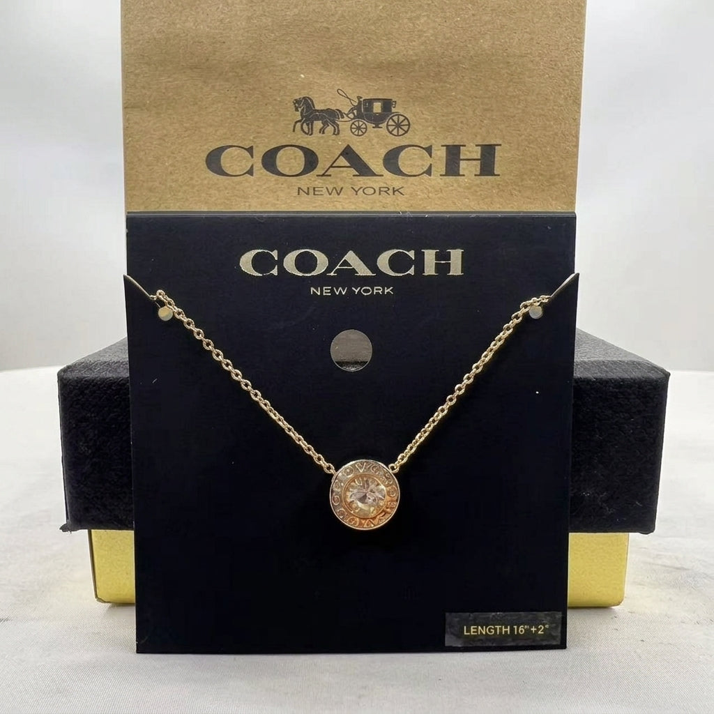 A gold-tone Coach New York necklace with a round pavé disc pendant, displayed on its black and gold branded retail packaging card.