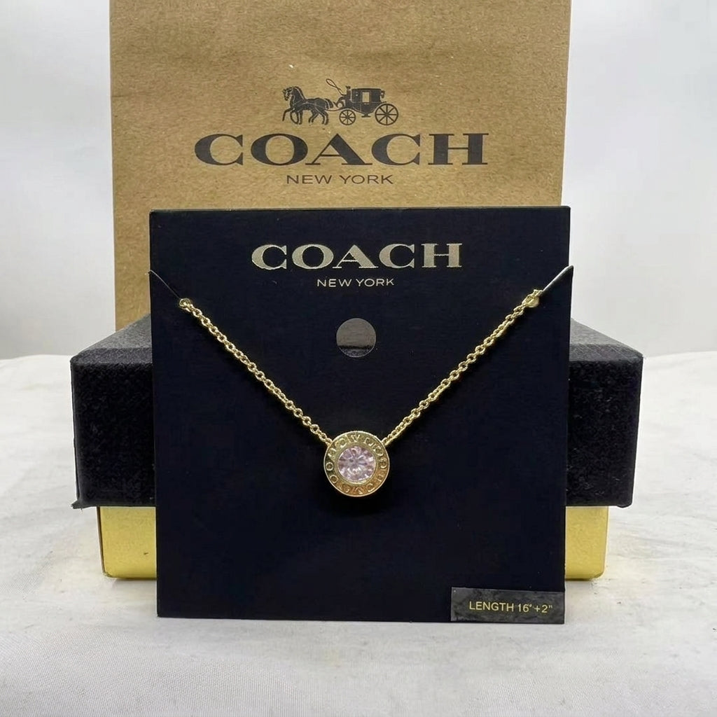 A gold-tone Coach necklace with a round pavé-set clear stone pendant, displayed on a black Coach New York card with its signature kraft paper box behind it.