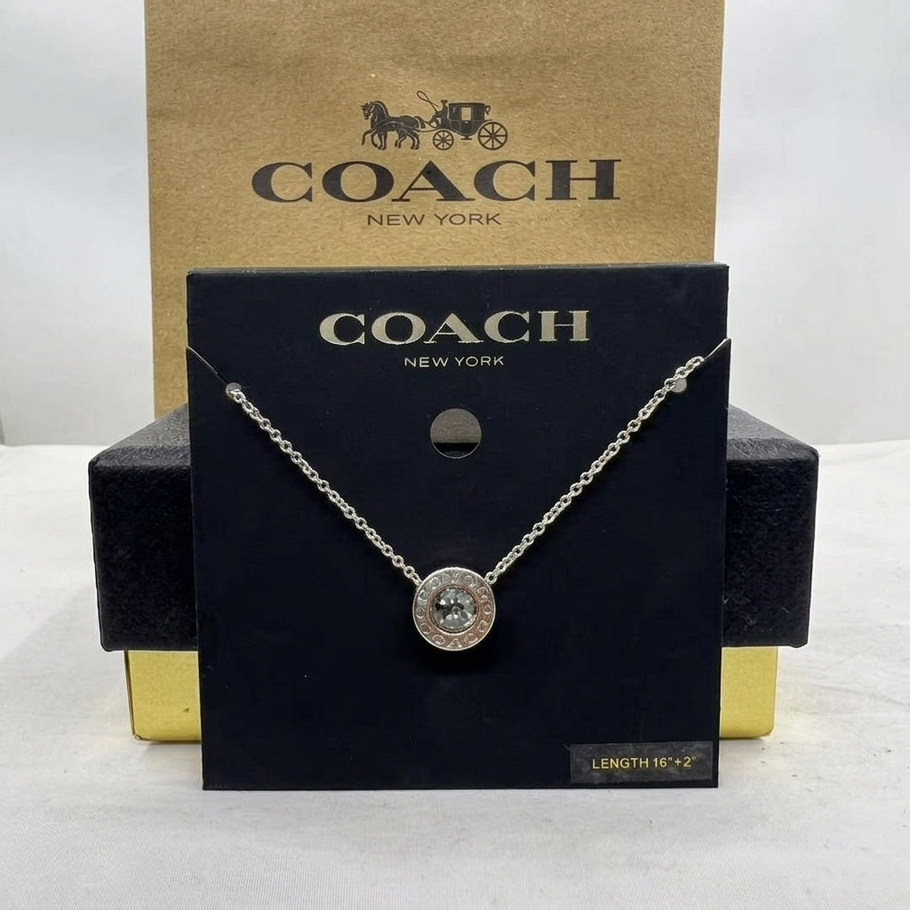 A Coach New York necklace displayed on a black branded card, with a gold Coach box behind it. The necklace has a silver-tone chain and a round pendant with a clear stone surrounded by a silver-tone border.