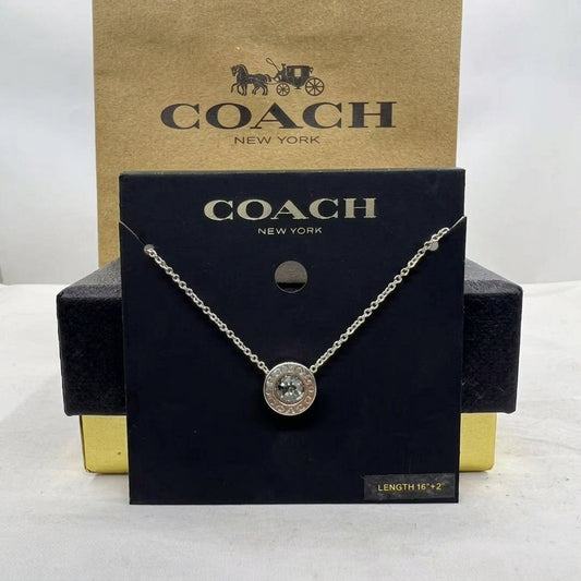 A Coach New York necklace displayed on a black branded card, with a gold Coach box behind it. The necklace has a silver-tone chain and a round pendant with a clear stone surrounded by a silver-tone border.