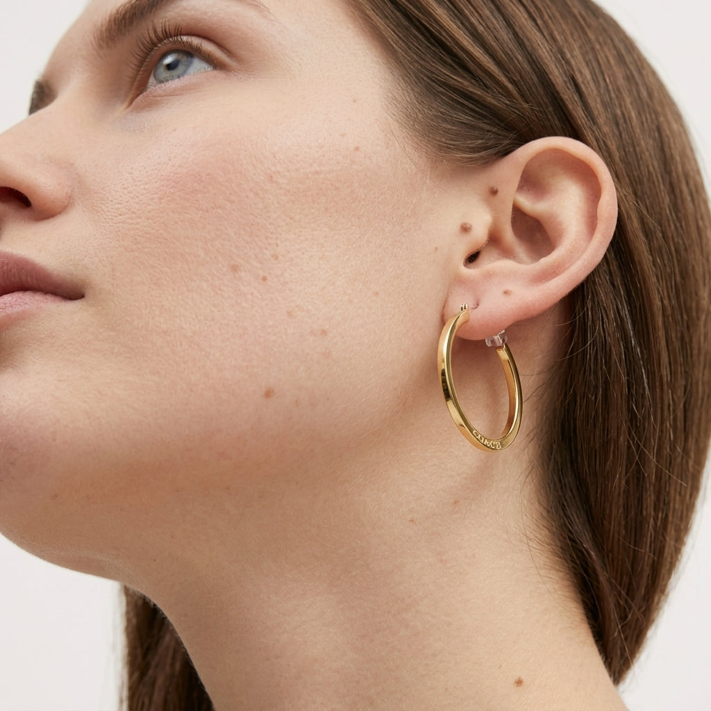 A close-up of a woman's ear wearing a polished gold hoop earring, with her face and neck shown against a clean light background.