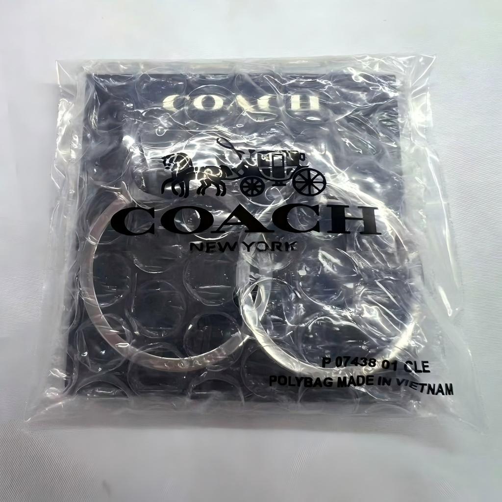 A Coach New York branded clear plastic polybag containing a pair of silver-toned metal key rings, photographed on a white background.