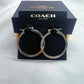 A pair of Coach silver-tone hoop earrings displayed on a black branded card in front of a dark Coach box on a white fabric background.