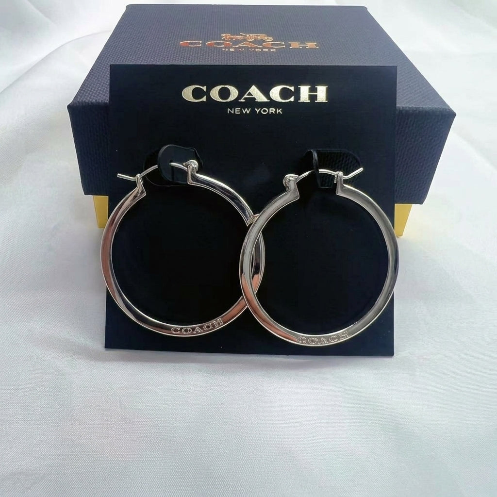 A pair of Coach silver-tone hoop earrings displayed on a black branded card in front of a dark Coach box on a white fabric background.