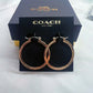 A pair of Coach hoop earrings in a polished rose gold finish, displayed on a black branded card in front of their dark navy blue presentation box.