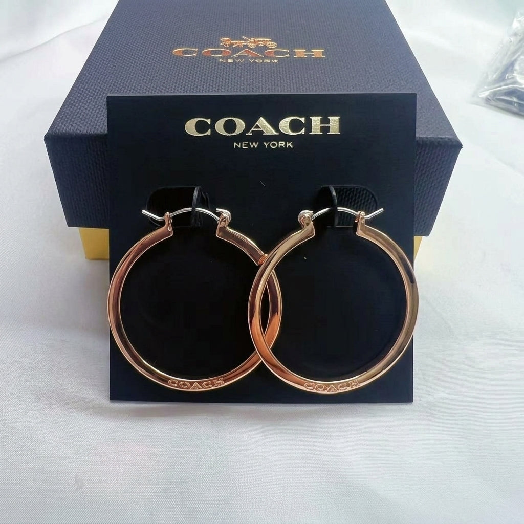 A pair of Coach hoop earrings in a polished rose gold finish, displayed on a black branded card in front of their dark navy blue presentation box.