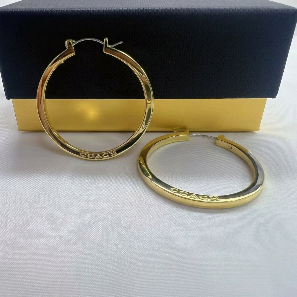 A pair of gold hoop earrings with the brand name 'COACH' engraved on the front, displayed in front of their black and gold presentation box on a white textured surface.