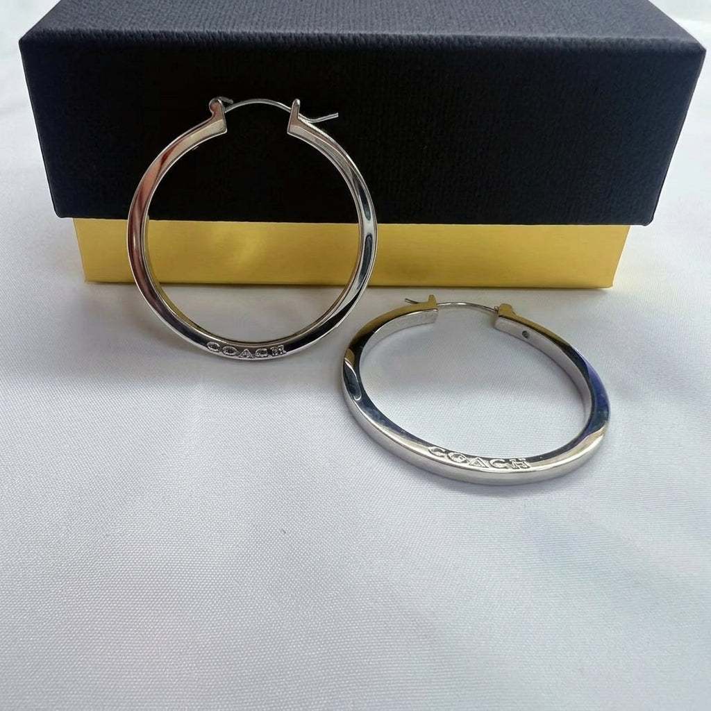 A pair of silver hoop earrings with 'COACH' engraved on the front, displayed in front of a black and gold Coach gift box on a white textured surface.