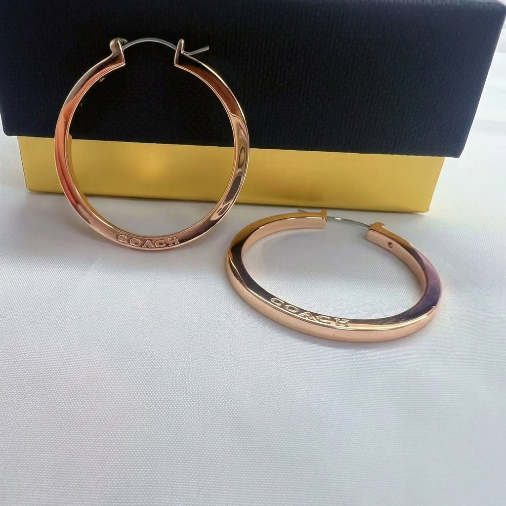 A pair of rose gold hoop earrings with the brand name 'COACH' engraved on the front, displayed in front of a black and gold presentation box on a white surface.