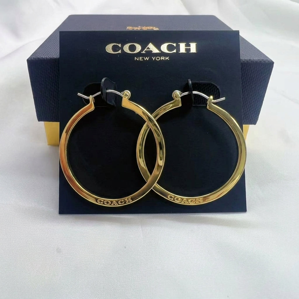 A pair of gold hoop earrings displayed on a black Coach New York card, with the branded navy box behind them on a white fabric background.