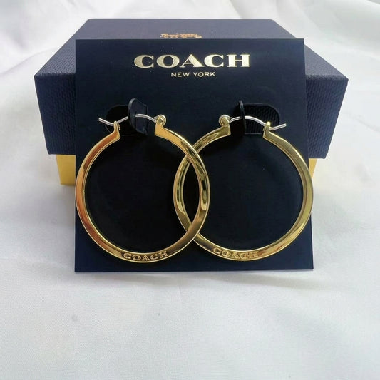 A pair of gold hoop earrings displayed on a black Coach New York card, with the branded navy box behind them on a white fabric background.