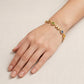 A gold bracelet with colorful gemstone and letter charms worn on a woman's wrist, shown against a light beige fabric background.