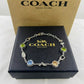A Coach New York bracelet displayed on a black branded box, with a tan Coach New York box behind it. The bracelet is silver-toned with multicolored gemstone charms and decorative links.