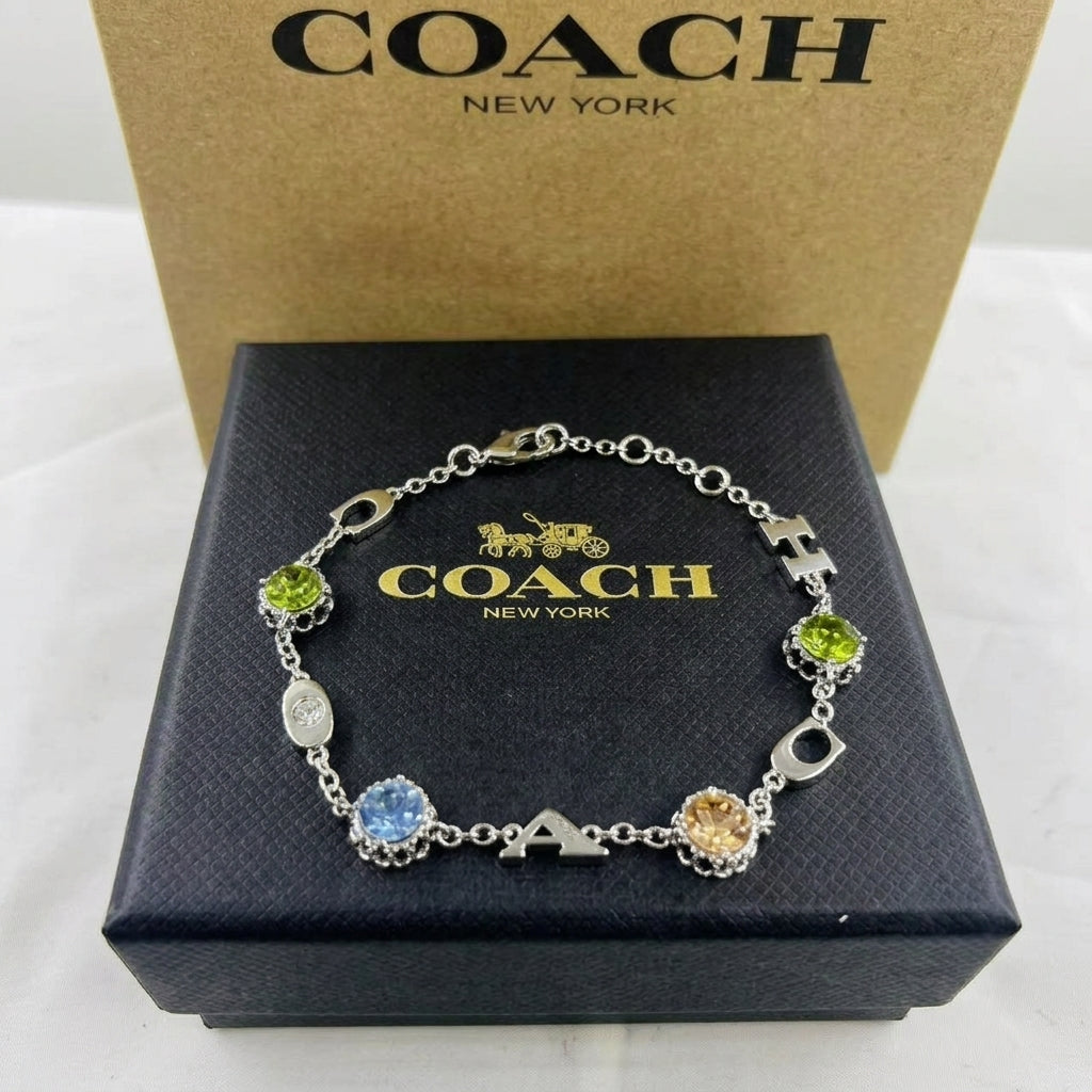 A Coach New York bracelet displayed on a black branded box, with a tan Coach New York box behind it. The bracelet is silver-toned with multicolored gemstone charms and decorative links.