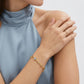 A close-up of a model wearing a delicate gold chain bracelet with a small light blue stone on her wrist, paired with a gold ring on her finger. She is wearing a high-neck dusty blue top against a clean, light gray background.