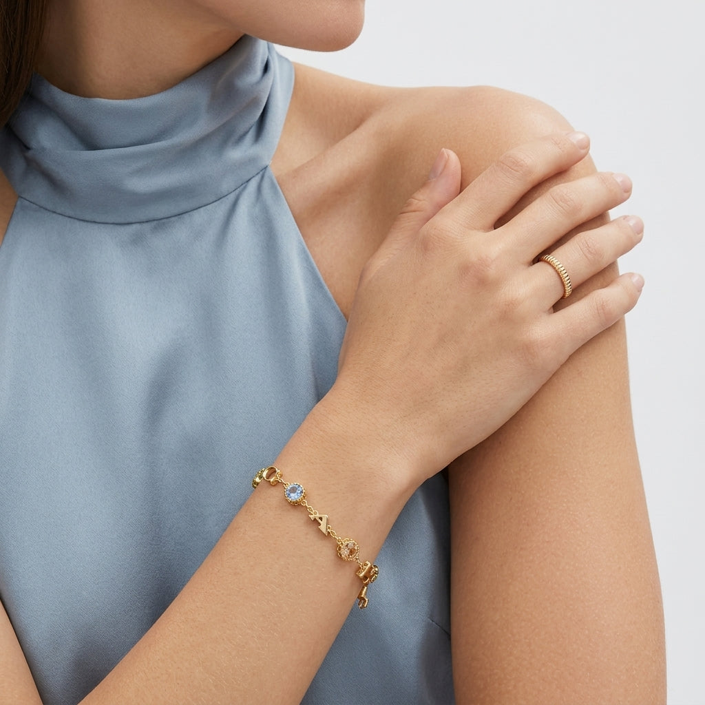 A close-up of a model wearing a delicate gold chain bracelet with a small light blue stone on her wrist, paired with a gold ring on her finger. She is wearing a high-neck dusty blue top against a clean, light gray background.