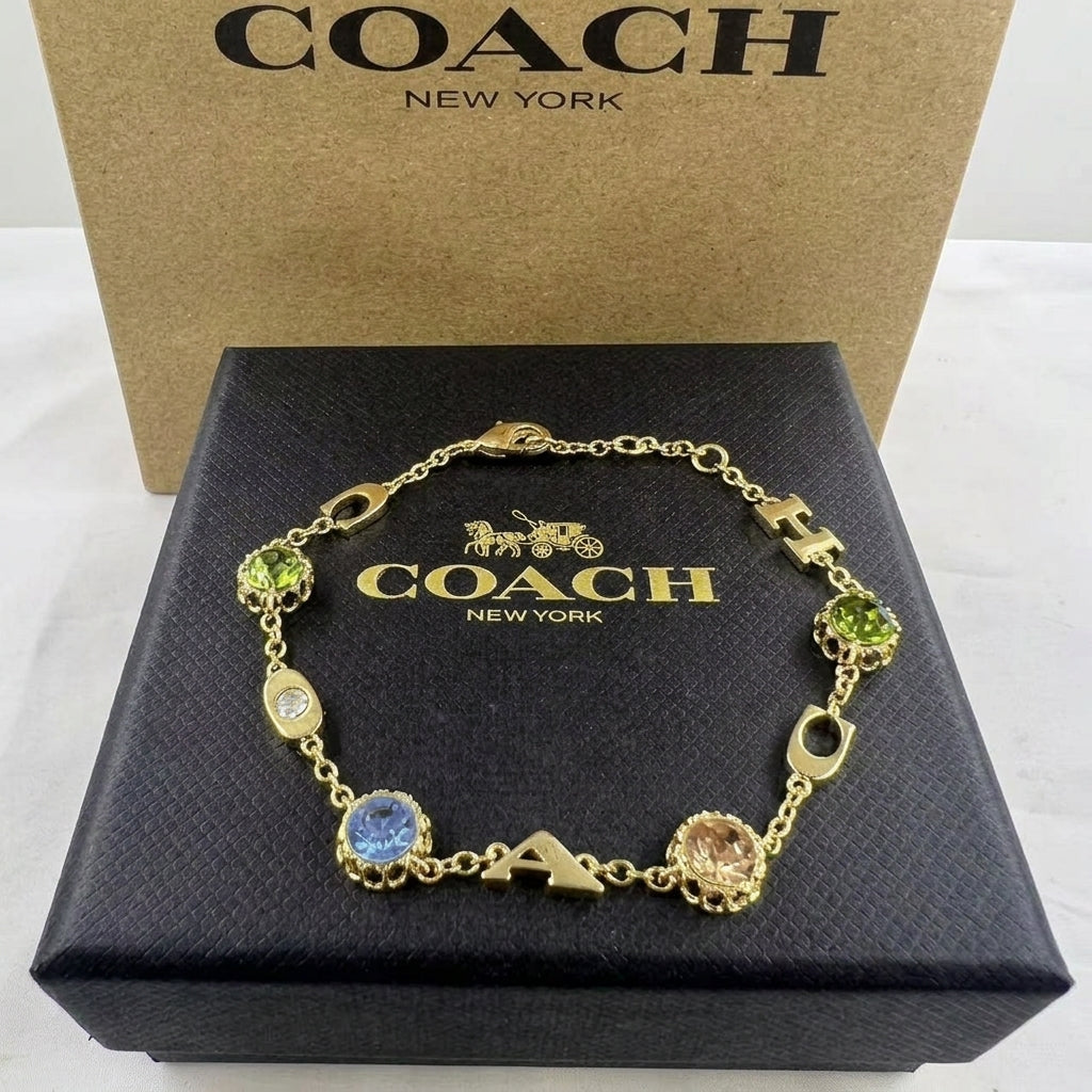 A gold-tone Coach letter charm bracelet displayed on its black presentation box, with the brown Coach New York box standing behind it.