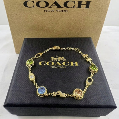 A gold-tone Coach letter charm bracelet displayed on its black presentation box, with the brown Coach New York box standing behind it.