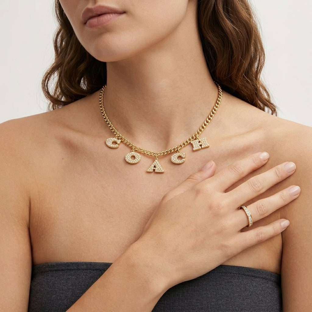 A woman wearing a gold-tone necklace with the word 'COACH' spelled out in letter charms, paired with a matching ring on her finger.