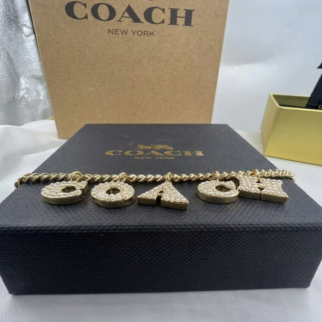 A gold-tone chain bracelet with four dangling letters 'COACH' covered in clear rhinestones, displayed on a black Coach box with a brown Coach box behind it.