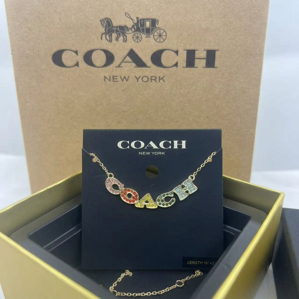A Coach letter necklace displayed on a black Coach New York card, with the gold-tone chain draped below. The necklace spells COACH using multicolored enamel letters on a gold base.