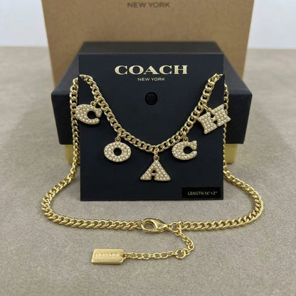 A gold-tone Coach letter charm necklace with rhinestone-embellished 'C', 'O', 'A', and 'X' charms, displayed on a black Coach New York box with a kraft box behind it.