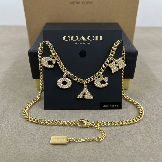 A gold-tone Coach letter charm necklace with rhinestone-embellished 'C', 'O', 'A', and 'X' charms, displayed on a black Coach New York box with a kraft box behind it.