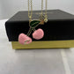 A gold-tone cherry pendant necklace with pink enamel cherries and a green leaf, displayed on top of a black and gold gift box.