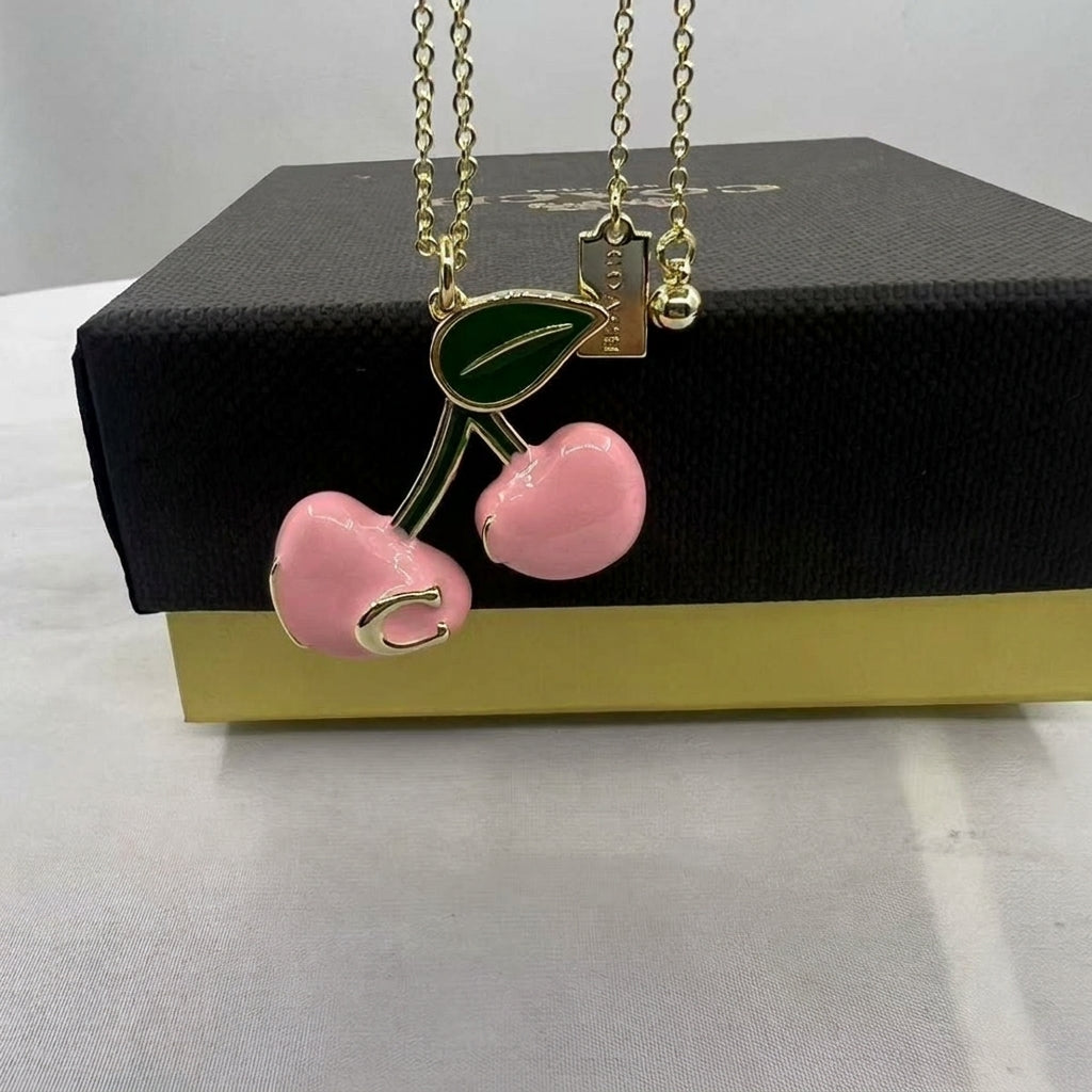 A gold-tone cherry pendant necklace with pink enamel cherries and a green leaf, displayed on top of a black and gold gift box.
