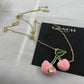 A gold-tone necklace with two pink cherry pendants and green leaves, displayed on a black Coach New York card against a white fabric background.