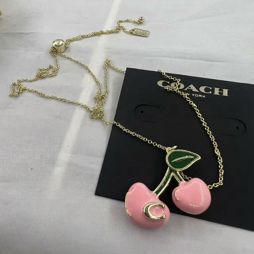 A gold-tone necklace with two pink cherry pendants and green leaves, displayed on a black Coach New York card against a white fabric background.