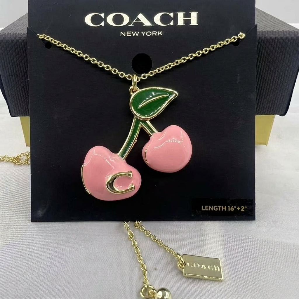 A Coach New York necklace featuring a cherry pendant with a pink body, green leaf, and gold-tone chain and lettering, displayed on a black branded card.