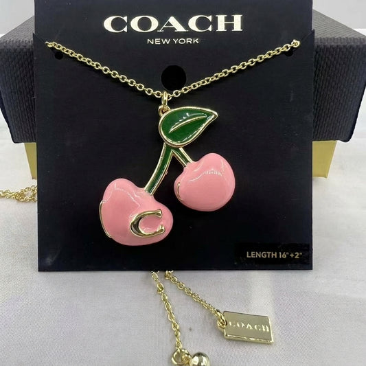 A Coach New York necklace featuring a cherry pendant with a pink body, green leaf, and gold-tone chain and lettering, displayed on a black branded card.