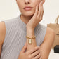 A close-up lifestyle shot of a model wearing a gold chain bracelet with a lock pendant. The model is wearing a light gray pleated sleeveless top, and a neutral background with a shopping bag is visible.