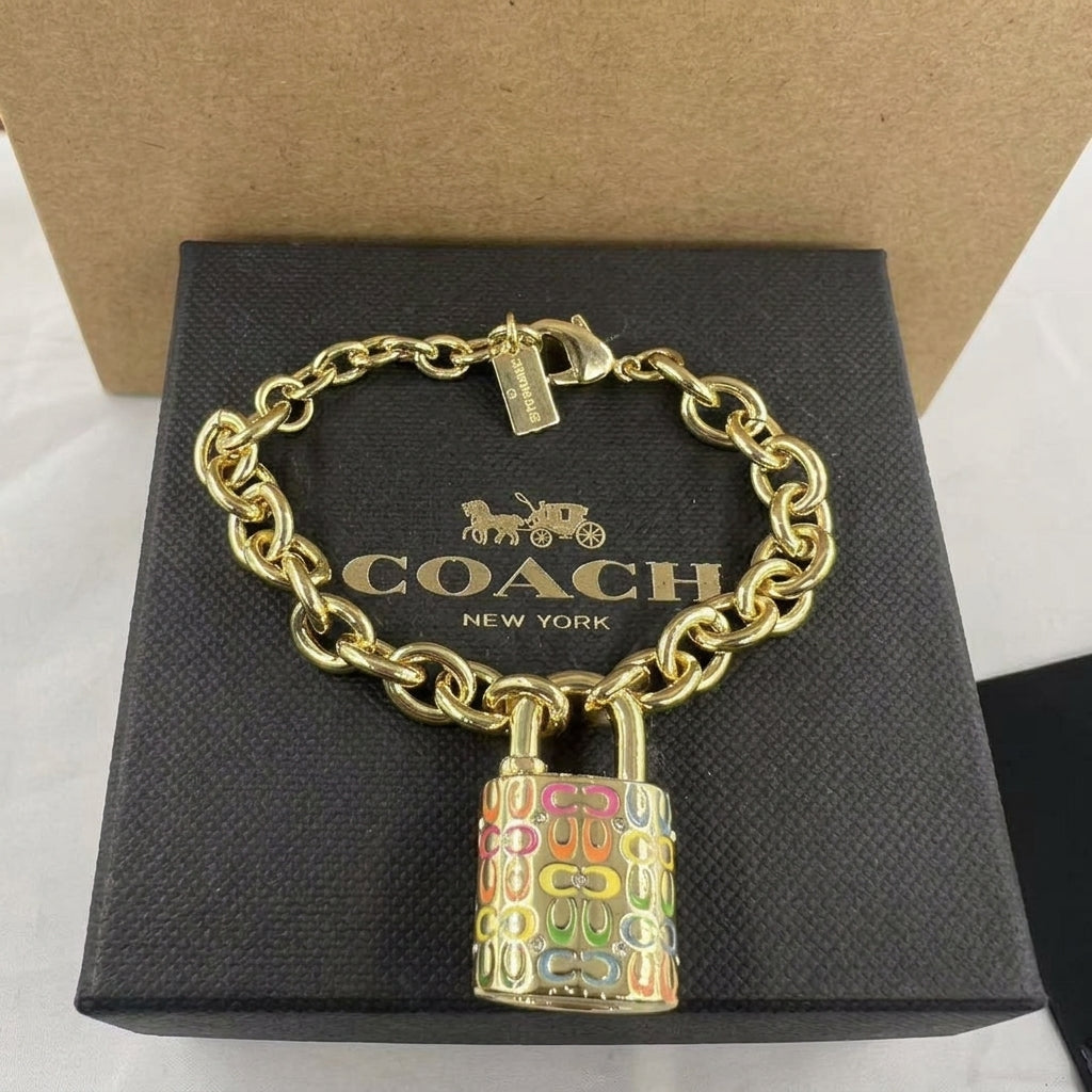 A Coach New York bracelet with a gold-tone chain and a colorful padlock charm, displayed on its black textured brand box.