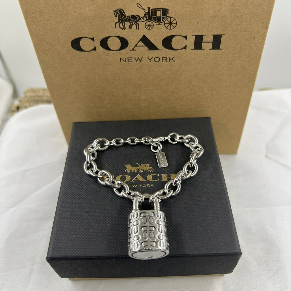A silver-tone chain bracelet with a padlock charm, displayed on a black Coach New York box with a brown Coach New York box behind it.