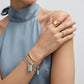 A close-up of a model wearing a stack of gold and silver bracelets on one wrist, including a chain bracelet with a padlock charm, alongside gold rings on her fingers, set against a light gray background and a dusty blue high-neck top.