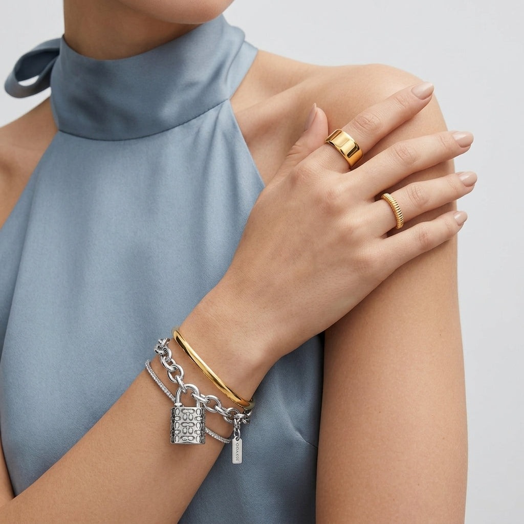 A close-up of a model wearing a stack of gold and silver bracelets on one wrist, including a chain bracelet with a padlock charm, alongside gold rings on her fingers, set against a light gray background and a dusty blue high-neck top.