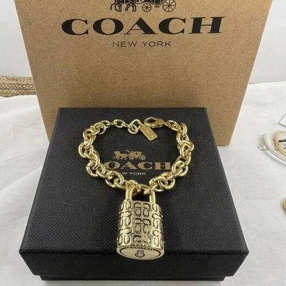 A gold-tone Coach chain bracelet with a padlock charm, displayed on a black Coach box in front of a brown Coach New York box.