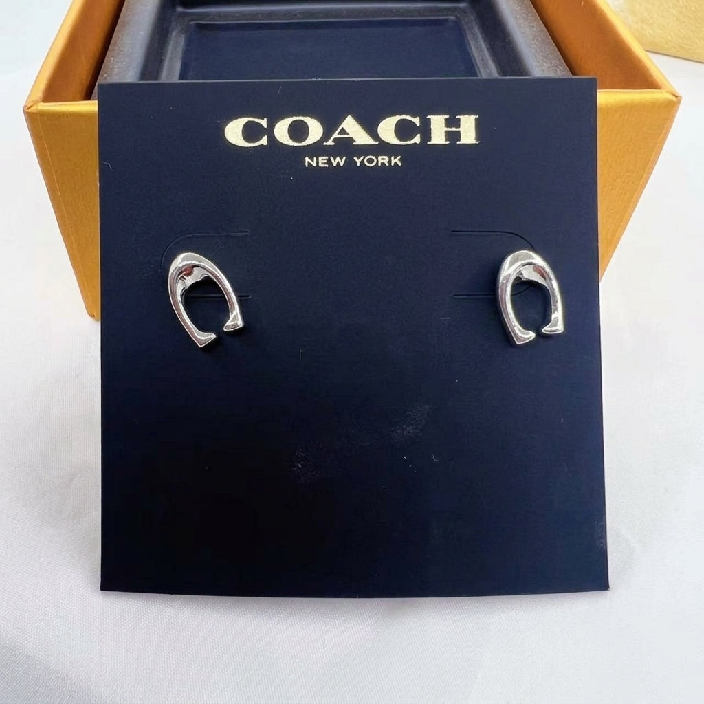 A pair of silver-tone Coach horseshoe hoop earrings displayed on a black Coach New York card insert, set against a gold and black Coach box on a white surface.