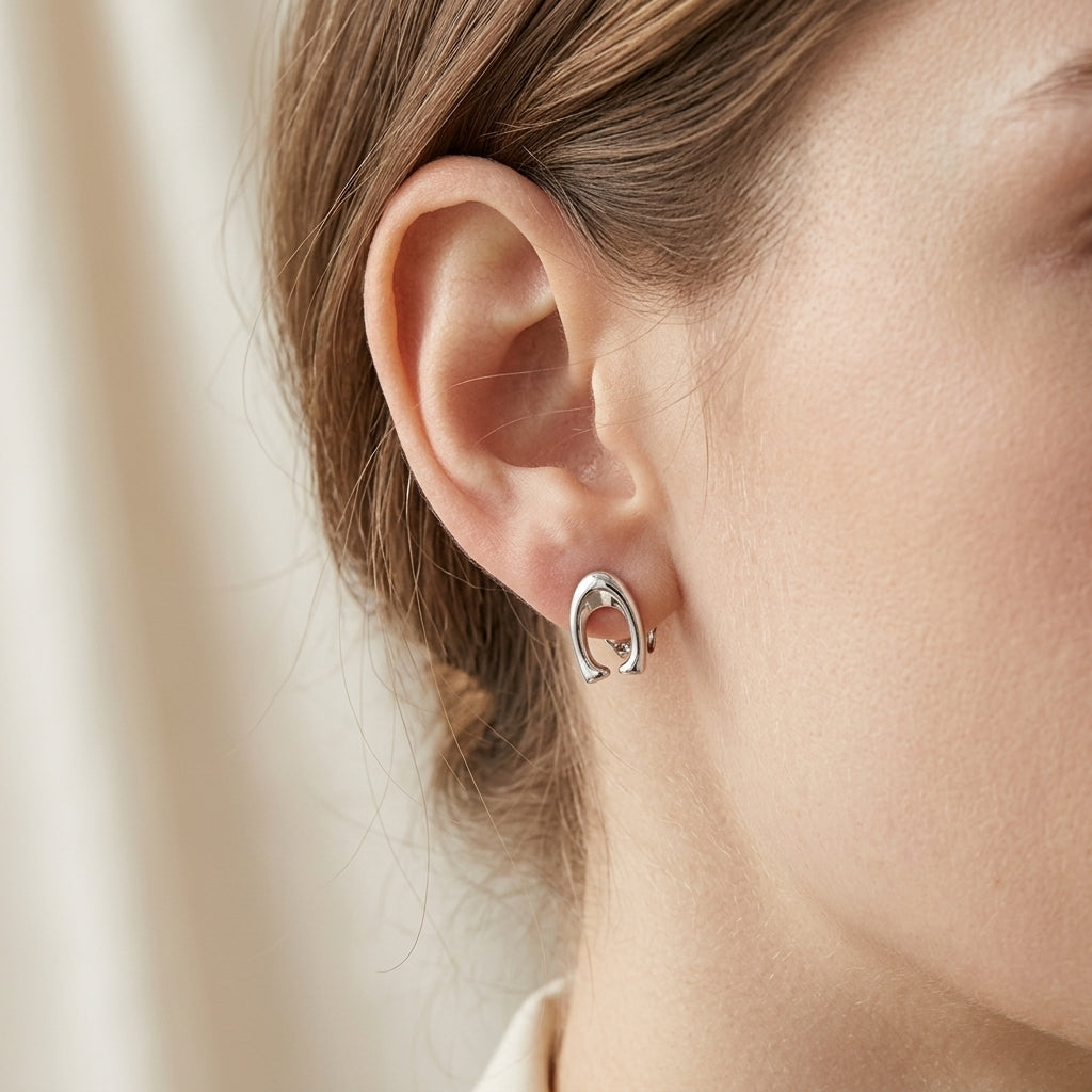 A close-up of a person's ear wearing a small silver hoop earring with a polished finish, set against a soft beige background.