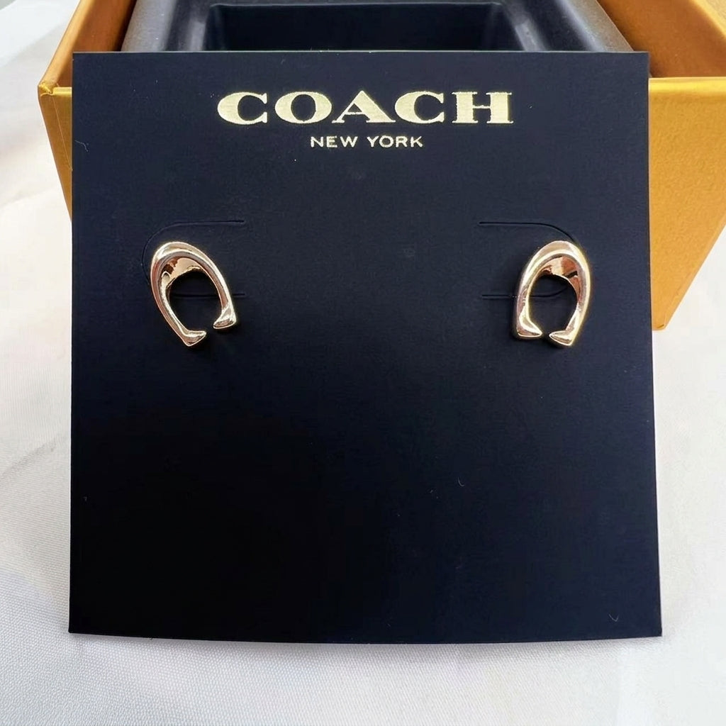 A pair of gold-tone Coach New York hoop earrings displayed on a black branded card, with part of a gold box visible behind it.
